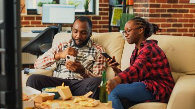 African american couple browsing apps on smartphones, using online internet on mobile phones and watching television together. Young modern partners using phones, takeaway food.