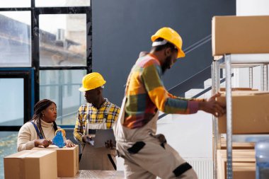 Depo çalışanları müşteri siparişi paketini yapıştırıcı ile paketliyor. Depodaki adam ve kadın çalışanlar karton kutu için yapışkan bant kullanıyorlar ve sipariş seçme biletini tartışıyorlar.