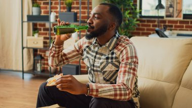 Young man eating slice of pizza at television on couch, enjoying takeout meal and bottle of beer at home. Modern person serving takeaway dinner and alcohol, watching film on tv.
