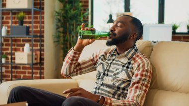 African american person eating burger and fries on sofa, having dinner with fast food and bottle of alcohol at home. Young man feeling happy watching action movie or show, having fun.