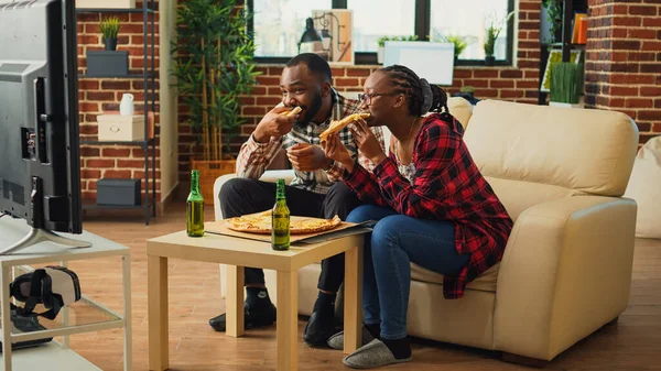 快乐的伴侣们在沙发上吃着披萨片 在家里欣赏着电视节目 享受着快餐餐 现代夫妇接受外卖食品和坐在电视机前吃饭 — 图库照片
