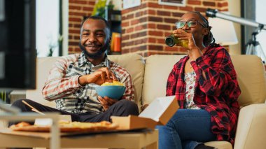 Young couple ordering takeaway food at home to watch television together, feeling relaxed with action film and bottles of beer. Man and woman laughing at tv show. Handheld shot.