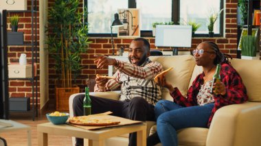 Happy couple eating slices of pizza at television, feeling relaxed watching movie together. Young life partners laughing and eating fast food delivery meal in living room, having fun.