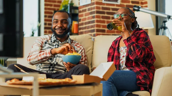 stock image Young couple ordering takeaway food at home to watch television together, feeling relaxed with action film and bottles of beer. Man and woman laughing at tv show. Handheld shot.