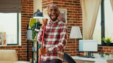 Modern happy boyfriend having fun with music while he uses mop to wash floors, mopping in apartment. Young male adult dancing and singing in living room, spring cleaning enjoyment.