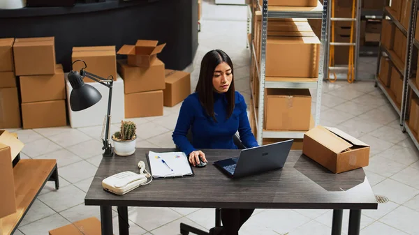 Small business owner planning distribution with goods, checking inventory and stock logistics for order shipment. Young woman working with laptop in storage room, financial planning.