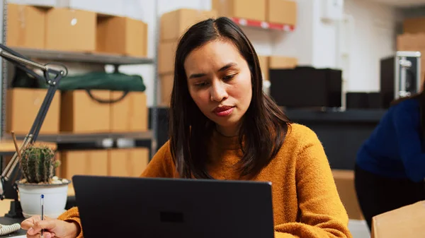 Young startup owner working on laptop to plan logistics, preparing products order to ship to clients. Female employee packing merchandise in boxes, taking notes for storage inventory.