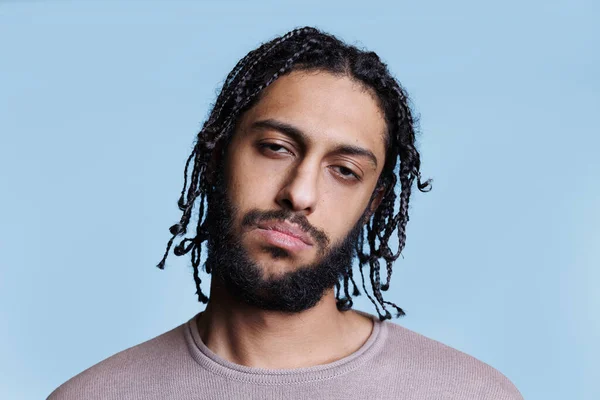 Cool confident attractive arab man tilting head with serious facial expression portrait. Young adult brunette arabian person with braids hairstyle and casual clothes looking at camera