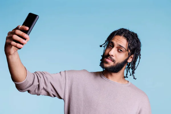 阿拉伯男子拿着智能手机 一边自拍一边看相机 穿着休闲装 在手机肖像上照相 在蓝色背景上摆姿势的年轻人 — 图库照片