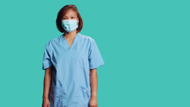 青いスタジオの背景に隔離された保護臨床面マスクを身に着けて幸せなアジアの看護師の肖像画 陽気Bipoc女性はプロの臨床スクラブの制服をスポーツ — ストック動画