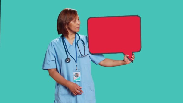 テキストのための空のスペースの赤いスピーチバブルサインを保持幸せな笑顔アジアの看護師 青いスタジオの背景に隔離された思考バブル段ボールを提示笑みを浮かべて医療専門家 — ストック動画