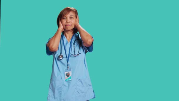 仕事中に過剰な騒音に悩まされる有資格看護師 耳を手で覆い 周囲の音に悩まされ 青いスタジオの背景に隔離された医療専門家 — ストック動画