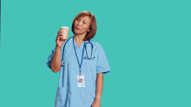 仕事の合間に淹れたてのコーヒーを楽しむ幸せなアジア系の有資格看護師 職場で熱い飲み物を飲みながらスタジオの背景に隔離された陽気な医療専門家 — ストック動画