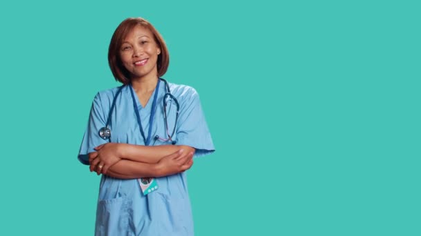 カメラを見ながら笑顔でアジアの看護師の折り畳み式の腕を 楽しい経験 青いスタジオの背景に隔離された保護スクラブを身に着けている幸せな医療専門家の肖像画 — ストック動画