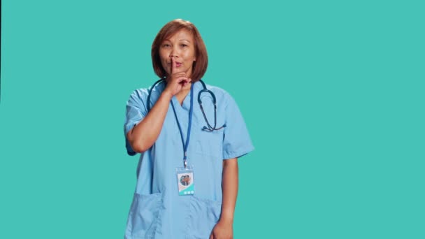 過度のノイズに悩まされ カメラで撮影された厳格な深刻なアジアの看護師の肖像画 青いスタジオの背景に隔離された唇に静かなジェスチャー指を保持する厳格なヘルスケアマネージャー — ストック動画