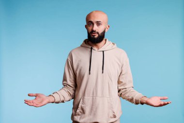 Arap adam şaşkınlık içinde duruyor, omuzlarını silkiyor ve belirsiz bir yüz ifadesiyle kameraya bakıyor. Gündelik kapüşonlu genç, yayılmış silah stüdyosu portresiyle.