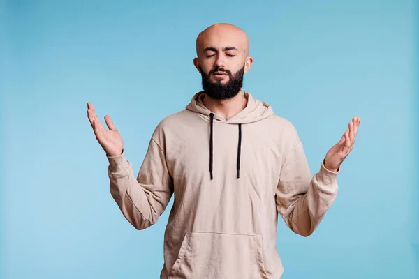 Arab man praying and making worship gesture with open arms and closed eyes. Young religious person praising, pleading to god with raised hands and asking for forgiveness concept