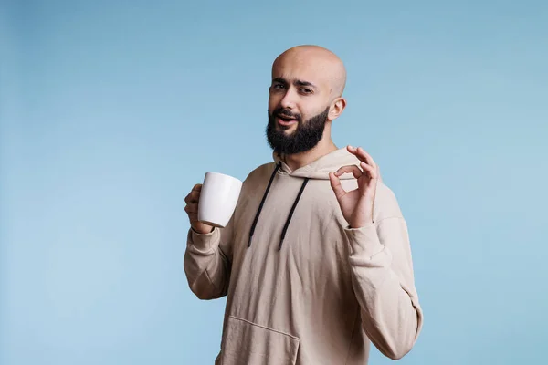 コーヒーを楽しみ 指の肖像画でOkジェスチャーを示すアラブ人男性 白マグカップでお茶を飲む若い人と承認サインと満足表情でカメラを見て — ストック写真