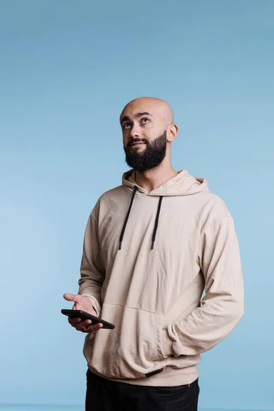 オンラインメッセージを入力し 上を見上げながら考えて スマートフォンを使用してアラブ人の男 パーカーのポケットに手で立っている間 携帯電話を保持している顔の表情を持つ若い人 — ストック写真