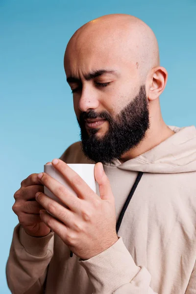 コーヒーを楽しみながら白いカップを手にしたリラックスしたアラブ人男性 青い背景にマグカップから熱いお茶を飲むカジュアルなパーカーを身に着けている若い思慮深い人 — ストック写真