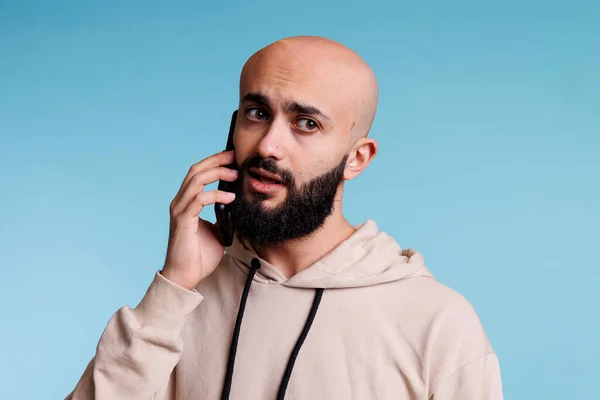 不确定的Arab男人在智能手机上说话 带着不确定的表情望着别处 年轻秃头的人在接听电话和交流时感到疑惑 — 图库照片