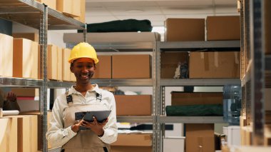 Afro-Amerikalı bir kadın, malzeme deposunda stok yapmak için tablet tutuyor, nakliye ürünlerini kontrol etmek için cihaz kullanıyor. Kadın işçi planlama düzeni dağılımı.