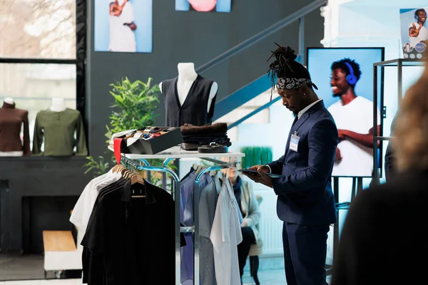 非裔美国人雇员在平板电脑上查看服装库存 在衣架上查看时尚商品 营业员在购物中心的设备上查打折单 — 图库照片