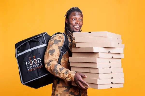 ピッツェリアの配達従業員は ピザでいっぱいの段ボールスタックを保持し ランチタイムの間に顧客にテイクアウト食品の注文を提供します スタジオで熱バックパックを運ぶアフリカ系アメリカ人の配達人 — ストック写真