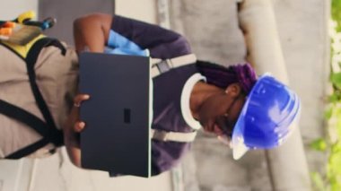 Dikey video deneyimli mühendis dış HVC sistemi üzerinde çalışıyor, dizüstü bilgisayar tutuyor. Açık hava yoğunlaştırıcı performansını optimize eden titiz bir kablocu maksimum kapasitede çalışmasını sağlıyor.