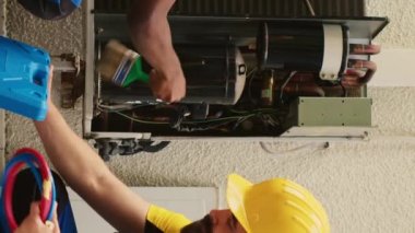 Dikey video bilgisi olan mühendisler klima içerisindeki sıvı ve gazları kontrol etmek için profesyonel basınç göstergesi oluştururken, Afro-Amerikan mekanikçi de toprak ve toz tabakasını temizliyor.