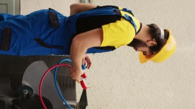 Dikey video teknisyeni havalandırmada soğutucu seviyelerini manifold göstergeleriyle ölçerken, Afrikalı Amerikalı iş arkadaşı çatı katlama merdivenini kontrol ettikten sonra merdivenden indi.