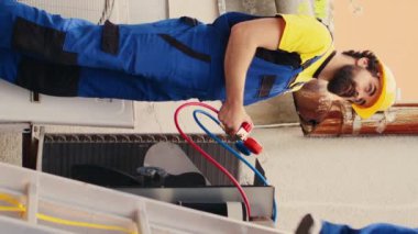 Meslektaşı soğutucuyu kontrol etmek için manometreyi kurarken, çatı hvac sistemini tamir ettikten sonra katlama merdiveninden inen elektrikli el matkabı tutan dikey video uzmanı