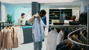 Afro-Amerikan müşteri alışveriş merkezindeki mağazada asılı duran resmi gömlek kumaşlarını inceliyor. Erkek müşteri modaya uygun ürünlere bakıyor, mağazadan modern kıyafetler alıyor..