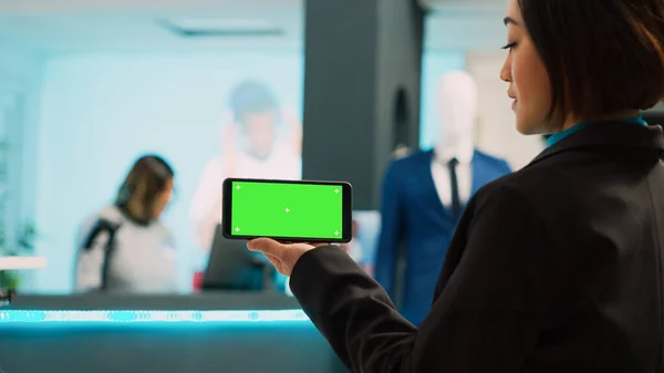 携帯電話に隔離された緑の画面を示すアジアのアシスタント クロマキーテンプレートとスマートフォンのディスプレイを使用して 女性従業員は 空白のモックアップとコピースペース 衣料品店でデバイスを保持 — ストック写真