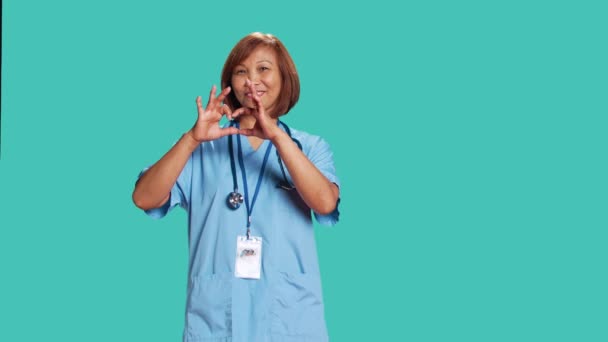 快乐的医疗保健专业人员用手做心形符号的肖像 有执照的亚洲护士在工作时表现出爱意 在蓝色工作室背景下被隔离 — 图库视频影像