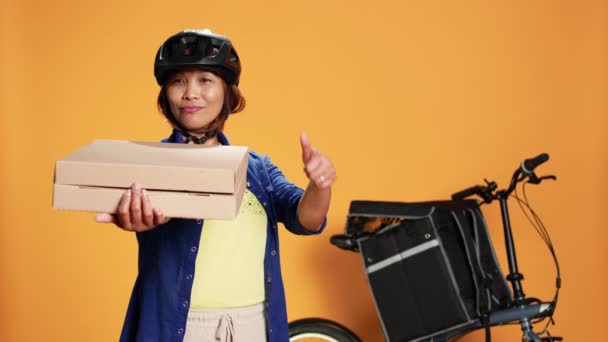 一个快乐的专业快递员指着外卖披萨盒的画像 在交付外卖食品时 微笑的骑自行车者竖起大拇指 在橙色工作室的背景下被隔离 — 图库视频影像