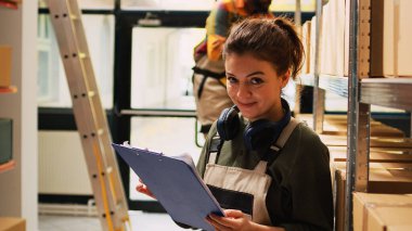 Depodaki kargo listesini inceleyen genç bir çalışan karton kutularla dolu rafları inceliyor. Tulum giyen kişi, ürün envanteri ve kağıt üzerindeki lojistik analizi, tedarik zinciri.