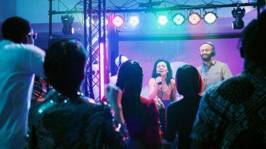 Kulüpte karaoke yapan gülümseyen kız, mikrofonda şarkı söyleyen ve sahnede DJ ile dans eden. Genç adam insanların eğlenmesi ve eğlenmesi için müziği karıştırıyor, dans partisi. El kamerasıyla..