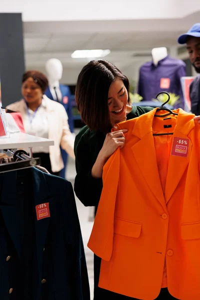 Zadowolona Azjatycka Kobieta Shopper Przymierza Klasyczną Pomarańczową Kurtkę Czerwoną Metką — Zdjęcie stockowe