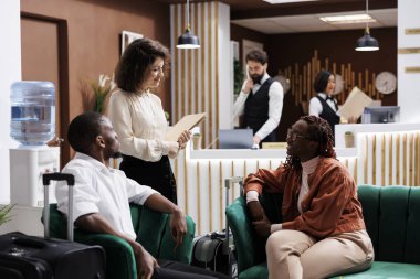 Yönetici misafirlere servisi soruyor, sıcak karşılama ve modern geniş odalar sağlamaya çalışıyor. Otel kapıcısı lobide insanlarla konuşuyor, personel konukseverliğini kontrol ediyor..