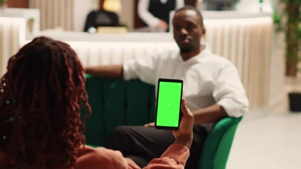 ホテルのロビーでチェックインされるのを待っている出張で会社のパートナーは クロマキー緑の画面モックアップスマートフォンを保持する 居心地の良いリゾートロビーでチャットする同僚 電話で情報を確認する — ストック動画