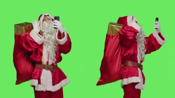 オンラインビデオコールで聖ニック スマートフォンのテレカンファレンスチャットで手を振ってプレゼントとバッグを保持します 携帯電話でのリモート会議を使用して クリスマスの精神を広める楽しいサンタキャラクター — ストック動画