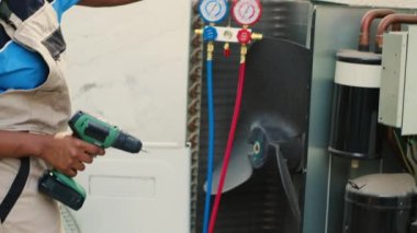 Nitelikli elektrikçi, kondansatör açmak ve manifold göstergelerini bağlamak için el matkabı kullanarak bozuk klima üzerinde çalışmaya başlıyor. İşçi içerideki basınç seviyelerini ölçmek için hvac sistemi açıyor