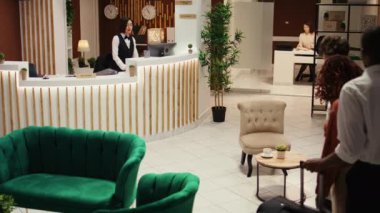 Dostça gülümseyen kapıcı personel otel lobisinde misafirleri karşılıyor. Profesyonel komi, turistlerin bagajlarını taşımalarına yardım ederken neşeli resepsiyonist de yardım çeki veriyor.