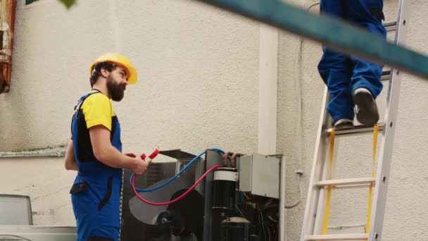有能力的电工用流量计测量冷凝器中的氟利昂电平 而非洲裔美国同事在检查完屋顶暖通空调系统后从折叠梯子上下来 — 图库视频影像