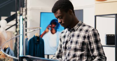 Yeni moda koleksiyonuna bakan Afrikalı Amerikalı alışverişçi modern butikte kumaş ürünlerini analiz ediyor. Alışverişkolik müşteri giyim mağazasından şık kıyafetler alıyor.