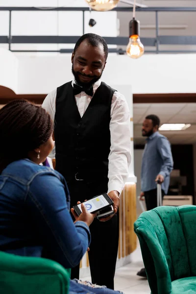 アフリカ系アメリカ人の男性ウェイター笑顔ロビーに座っている女性のホテルのゲストにPossマシンを与える ラウンジエリアでチェックイン手続きを待っている間 スマートフォンを持っている観光客は注文を支払う — ストック写真