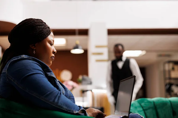 认真关注的非洲裔美国女性旅行者坐在酒店大堂时凝视着笔记本电脑屏幕 女性客人使用电脑上网登记 远程工作和旅行概念 — 图库照片