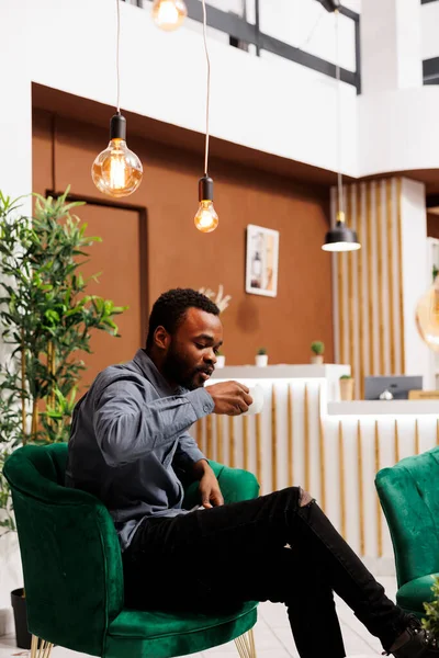 若いアフリカ系アメリカ人ビジネスマンがホテルのロビーでコーヒーを飲みながら ビジネスパートナーを待っているラウンジエリアで休んでいます 出張中にエスプレッソを楽しむ起業家 — ストック写真