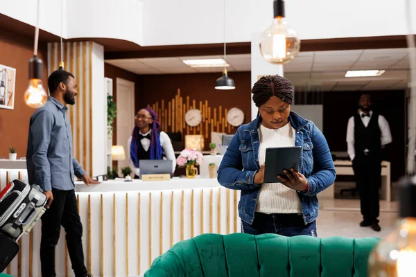 非洲裔美国妇女游客站在酒店大堂 手里拿着数字平板电脑进行自助登记 使用电子设备完成登记程序的女旅客 网上订票室 — 图库照片
